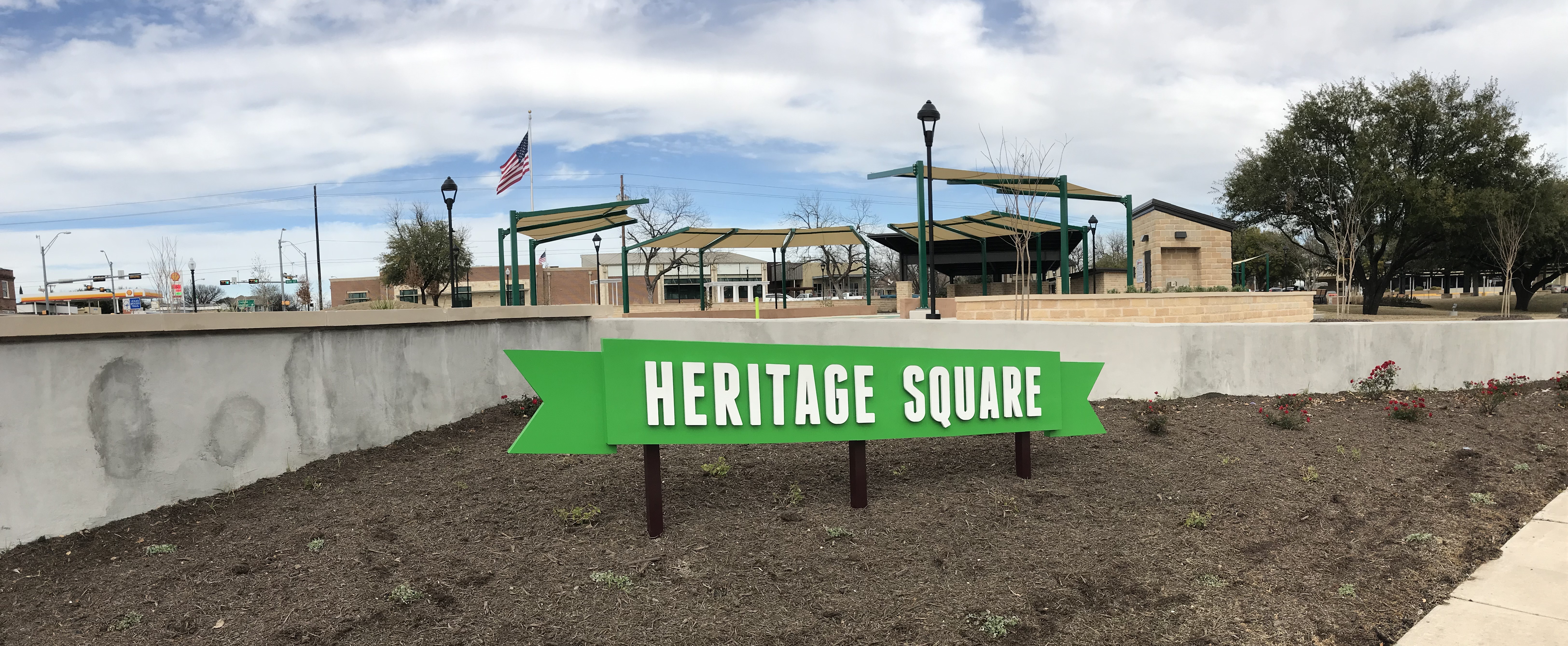 Green Heritage Square sign with park shade structures behind it.